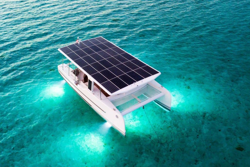 A solar catamaran at anchor in turquoise water on the Costa Brava, seen from above in low sun